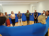 Teilnehmerinnen des Assistent*innen Forums stehen in einem Kreis, um sie herum ein blaues Tuch gespannt