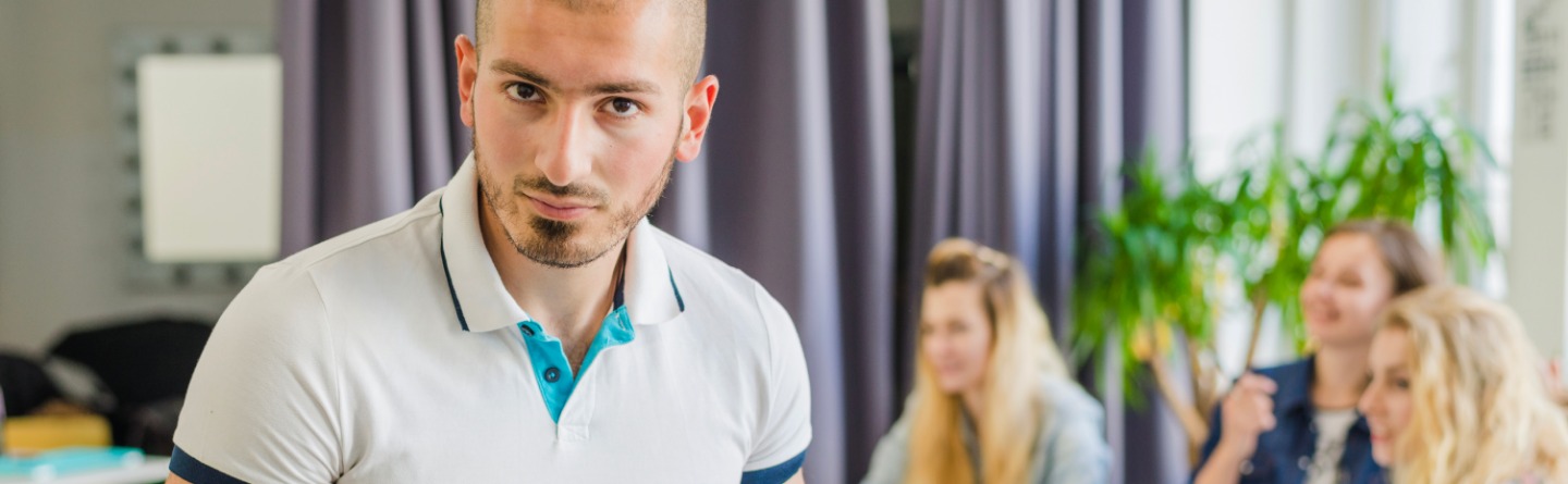 Junger Mann sitzt mit Blick in die Kamera auf einem Tisch, im Hintergrund sitzen junge Menschen am Tisch mit Lernutensilien