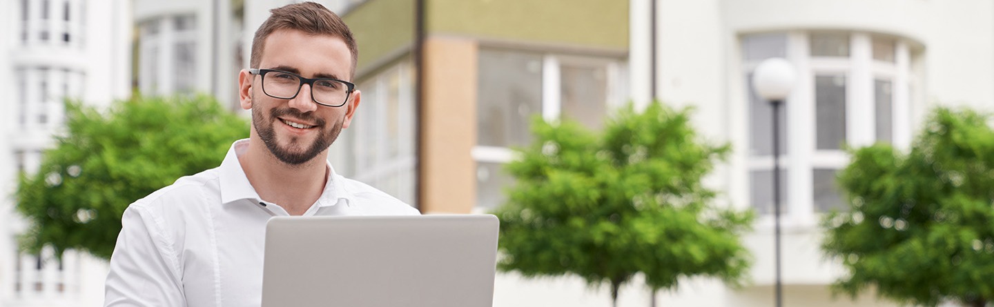 Junger Mann mit weißem Hemd, Brille und Notebook lächelt in die Kamera.