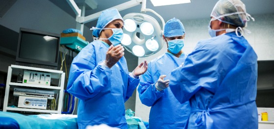 Drei Menschen stehen in einem Operationssaal und haben OP-Bekleidung an und bereiten sich auf die Operation vor.