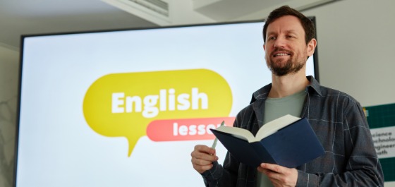 Ein Mann steht mit einem aufgeschlagenen Buch in der Hand vor einem Monitor auf dem "English Lession" geschrieben steht.