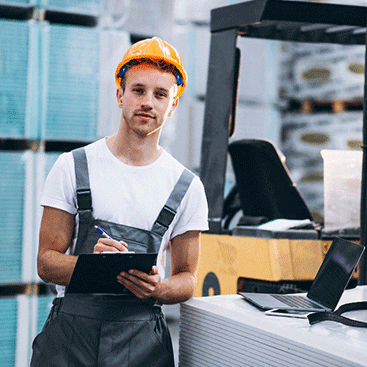 Ein junger Arbeiter im Overall und mit Schutzhelm steht vor eine Gabelstapler und hat ein schwarzes Klemmbrett in der Hand