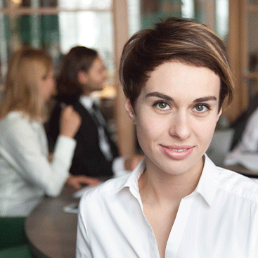 Eine Frau in weißer Bluse lächelt in die Kamera während ihre Kolleg*innen sich am Tisch im Hintergrund unterhalten