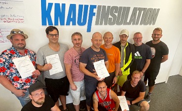Erfolgreiche Mitarbeiterschulung bei Knauf Gruppenbild nach einer Mitarbeiterschulung vor dem Firmenschriftzug Knauf Insulation. Mehrere Personen halten Teilnahmeurkunden in der Hand.