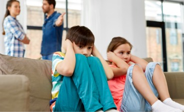 2 traurige Kinder auf einer Couch mit streitenden Eltern im Hinergrund