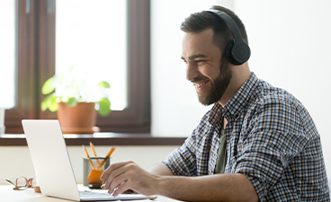 Ein junger Mann mit Bart und Kopfhörern sitzt am Schreibtisch vor seinem Laptop und lächelt in die Kamera.
