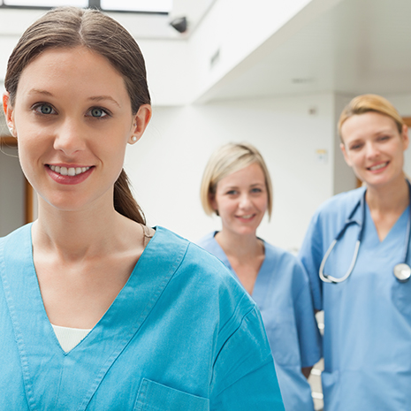 Eine Pflegerin steht im Vordergrund und lächelt in die Kamera während zwei ihrer Kolleginnen im Hintergrund stehen und auch in die Kamera lächeln