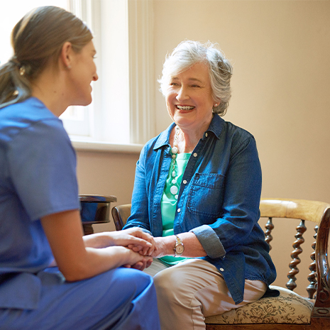 Eine Pflegerin sitzt einer Seniorin gegenüber und hält ihre Hand während sie sich anlächeln.