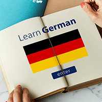Ein offenes Buch liegt auf einem Tisch während zwei Hände die Ecken des Buches halten. Auf den beiden Seiten steht "Learn German" und darunter ist die deutsche Flagge abgedruckt.